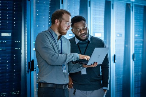 Bearded IT Technician in Glasses with Laptop Computer and Black Male Engineer Colleague are Using Laptop in Data Center while Working Next to Server Racks. Running Diagnostics or Doing Maintenance Work