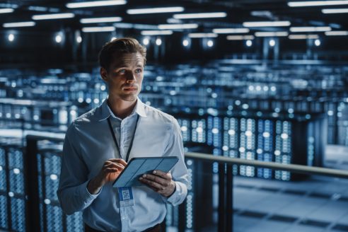 Portrait of Smiling IT Specialist Using Tablet Computer in Data Center. Big Server Farm Cloud Computing Facility with Male Maintenance Administrator Working. Cyber Security, e-Business.